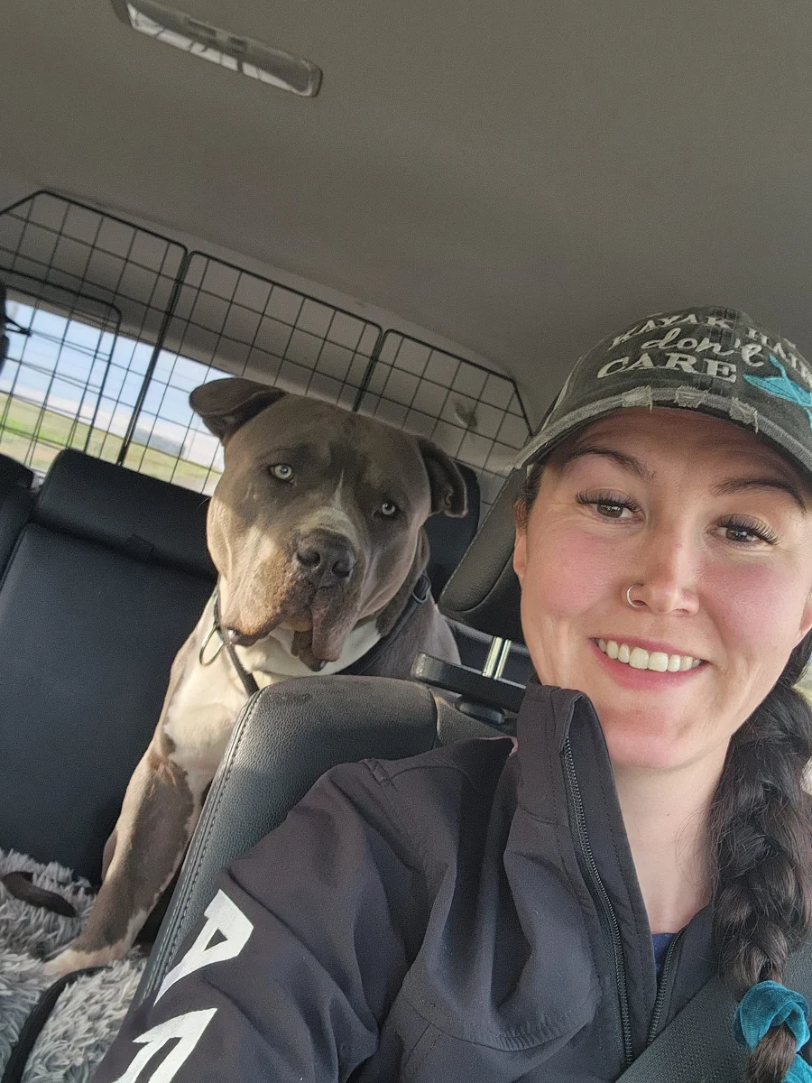 Dezeray Zellmer smiling in a cap in her car, with a grey pit bull in the back seat.